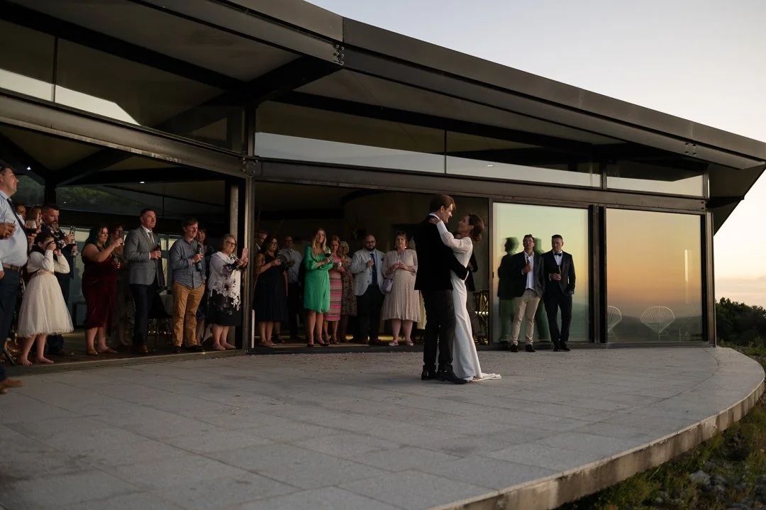 A bride and groom sharing a dance on a curved outdoor stone patio during sunset, with wedding guests watching behind them in front of a modern building with large glass windows.