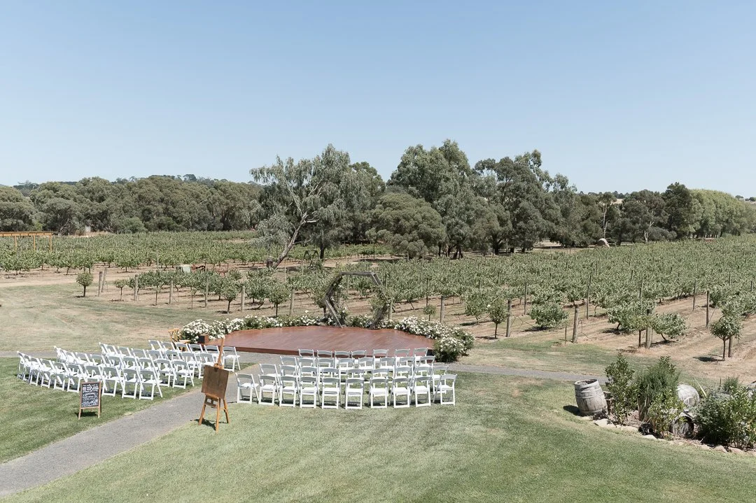 Outdoor wedding ceremony setup with white chairs arranged in front of a small stage with flowers, overlooking a vineyard with rows of grapevines and trees under a clear blue sky.