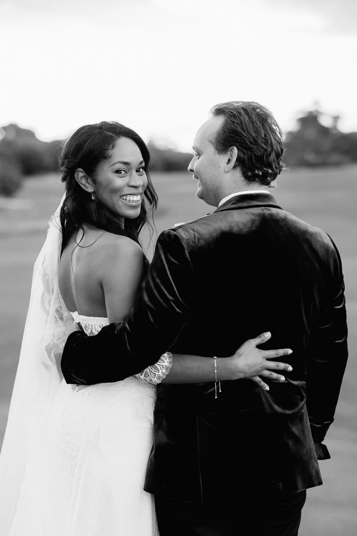 A smiling bride with dark hair and earrings, wearing a white wedding dress, embracing a groom with light hair and a beard, dressed in a black suit, outdoors on a grassy field.