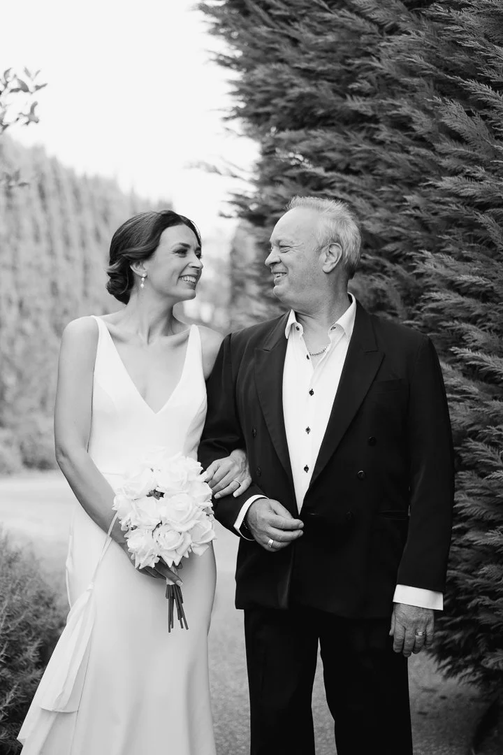 A bride and an older man, likely her father, smiling and looking at each other outdoors during a wedding. The bride is holding a bouquet of flowers.