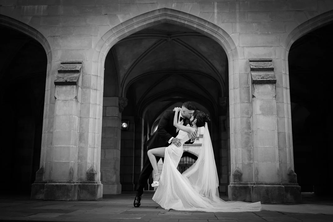 A wedding couple in formal attire sharing a kiss under arched stone architecture, with the groom dipping the bride in black and white.