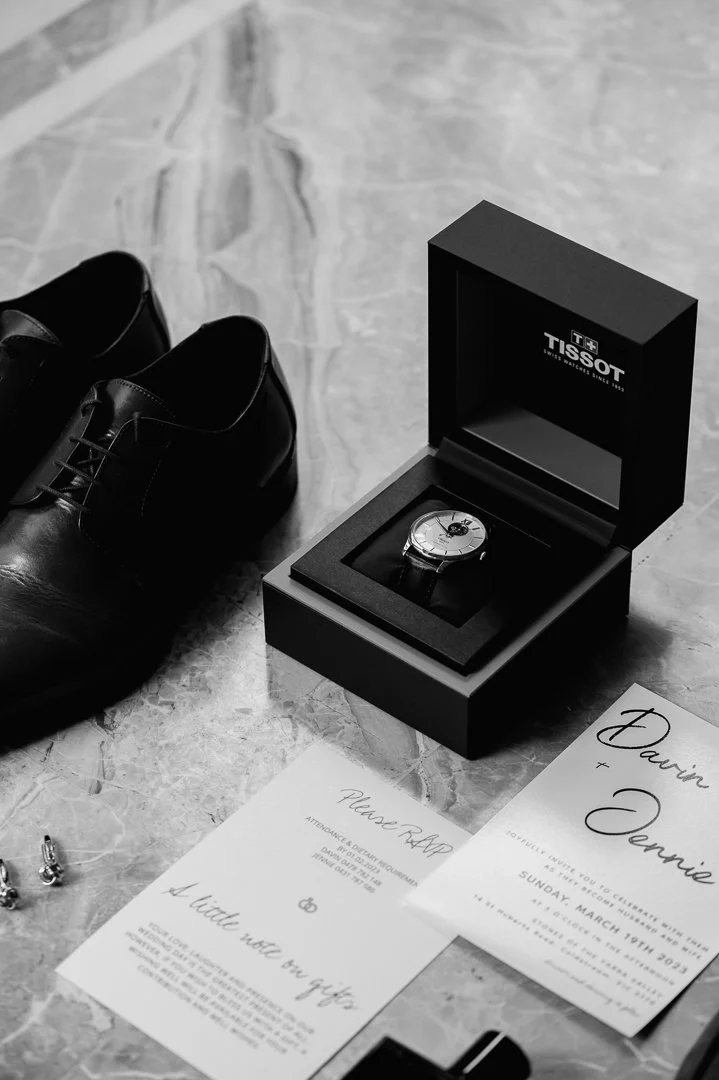 Black men's dress shoes, a Tissot wristwatch in its box, and wedding invitations on a marble surface.