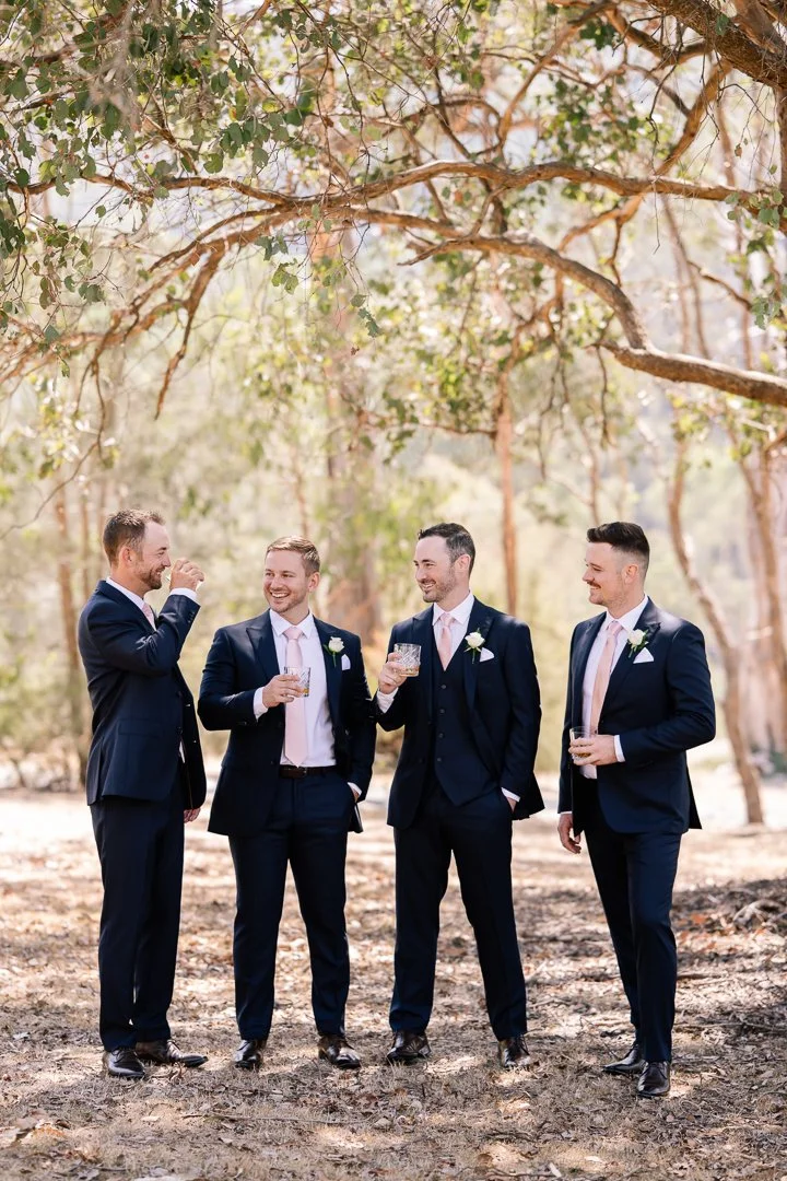 Four men in suits with pink ties holding drinks, standing outdoors under tree branches, engaged in conversation and smiling.