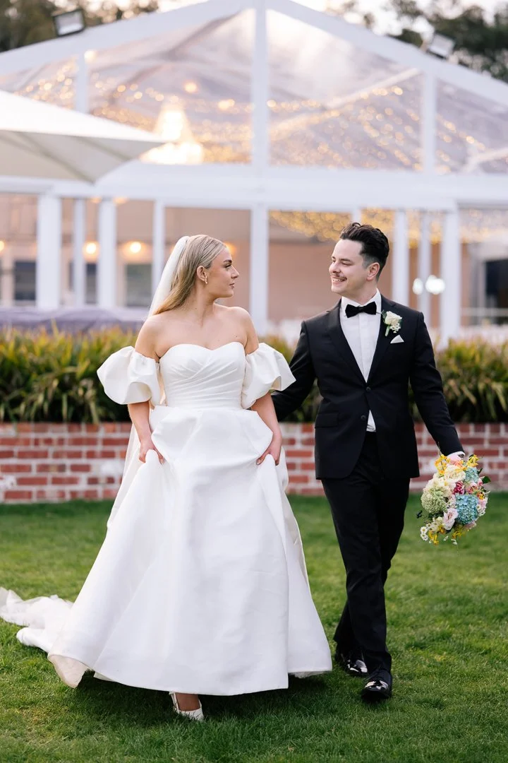 Bride and groom walking outdoors during wedding, bride in white gown with puff sleeves, groom in black tuxedo holding bouquet, festive string lights overhead, brick wall and greenery in background.