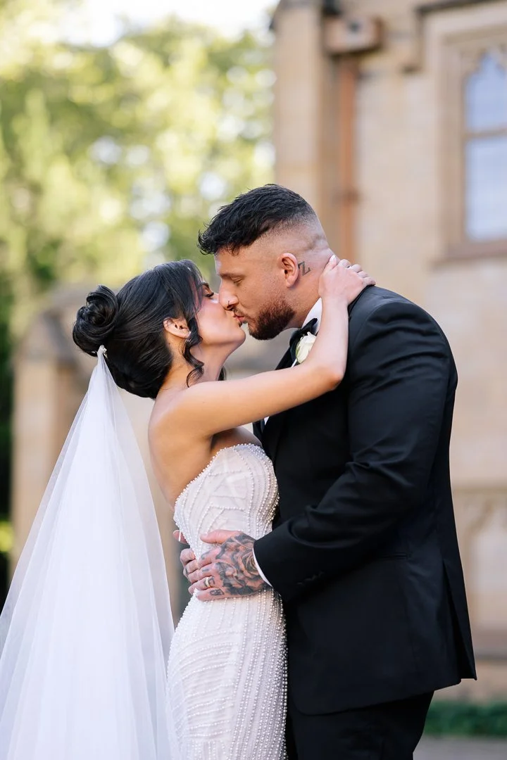 A bride and groom sharing a kiss outdoors during their wedding, the bride in a white gown with a veil and the groom in a black tuxedo.