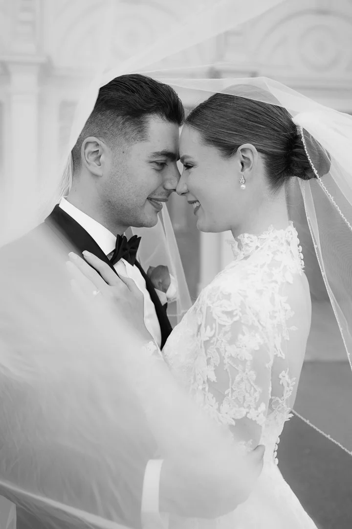 A black and white photo of a bride and groom under a sheer veil, smiling and touching foreheads, in wedding attire.