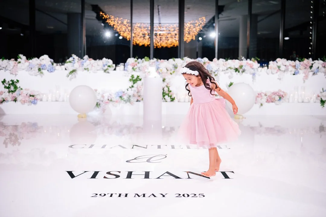 A young girl in a pink dress and headband dancing or spinning indoors in front of a floral backdrop and decorative lighting, with text on the floor reading 'Chantelle & Vishant 29th May 2025'