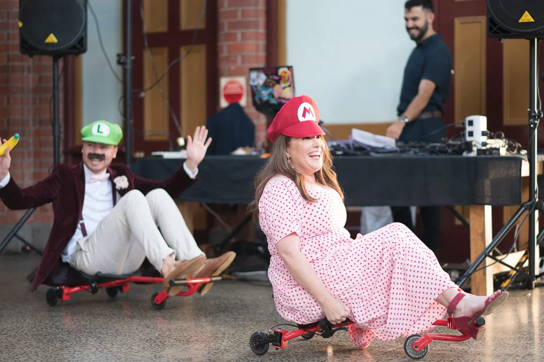 Two people dressed as Luigi and Mario from Super Mario riding small scooters indoors at a party. Luigi is wearing a green hat and moustache, while Mario is wearing a red hat and a polka dot dress. They are smiling and in the background, a DJ and man