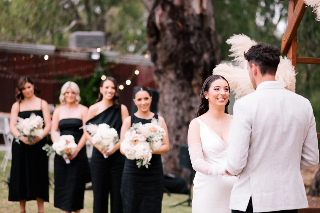 A bride and groom exchange vows outdoors while four bridesmaids stand in the background holding bouquets.