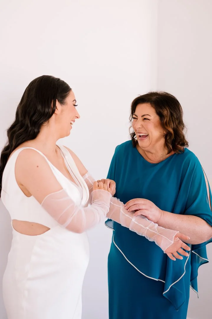 A woman in a white dress with sheer sleeves smiling and holding hands with an older woman in a blue dress, both sharing a joyful moment.