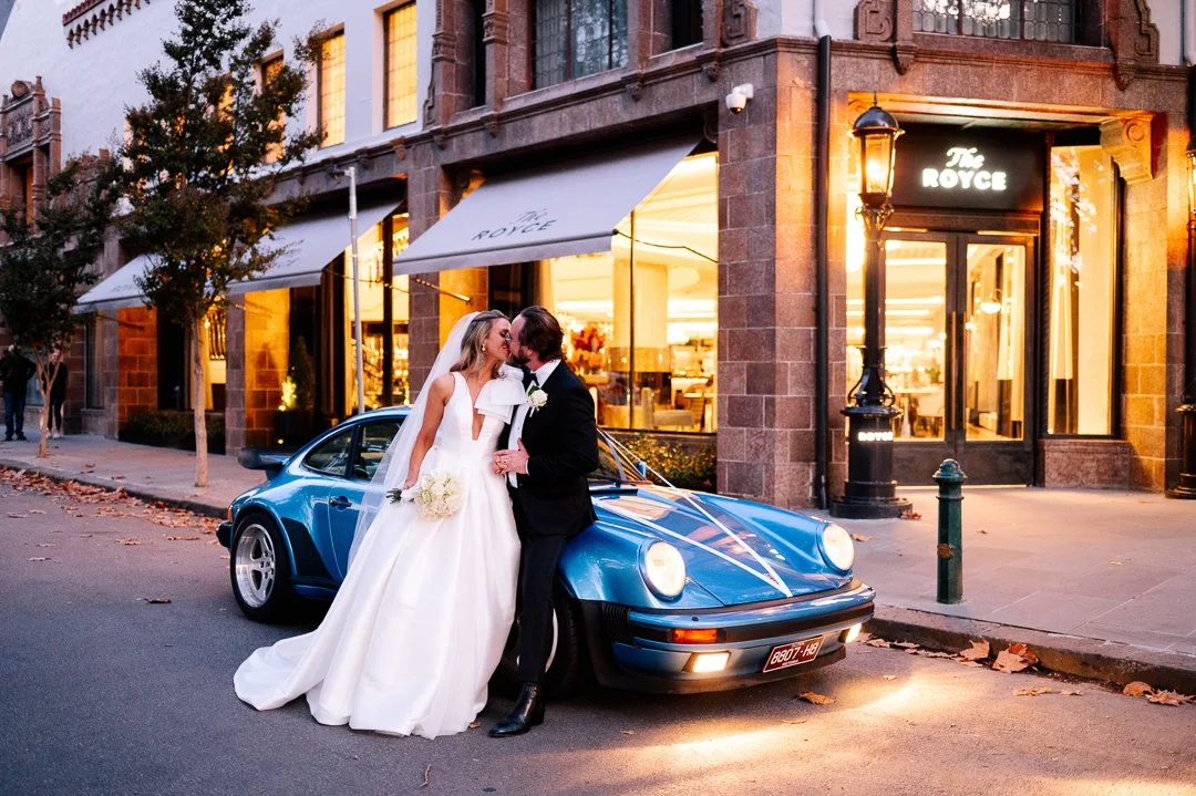 Bride and groom in wedding attire sharing a kiss beside a blue vintage sports car parked on a city street at sunset.