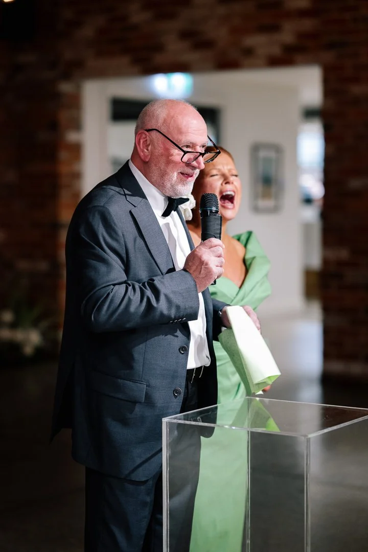 An elderly man in a tuxedo and glasses speaking into a microphone while a woman in a green dress laughs in the background at a formal event.