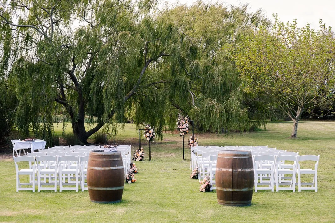 Outdoor wedding ceremony setup with white chairs, floral arrangements, and wooden barrels on a grassy lawn under large green trees.