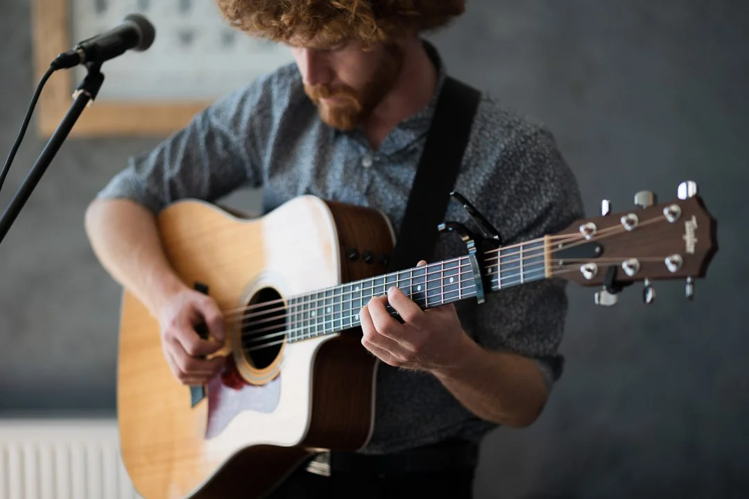 A man with curly hair and a beard playing an acoustic guitar in front of a microphone.