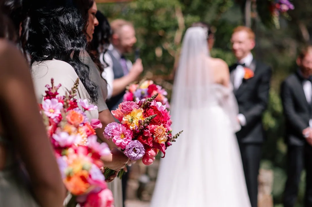 A wedding ceremony outdoors with bridesmaids holding colorful floral bouquets and a bride and groom in the background, surrounded by greenery.