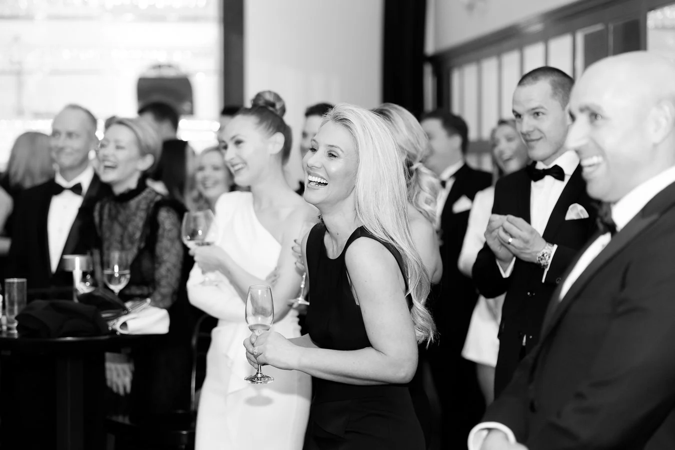 Guests at a formal event, dressed in tuxedos and elegant dresses, smiling and holding glasses of wine or champagne.