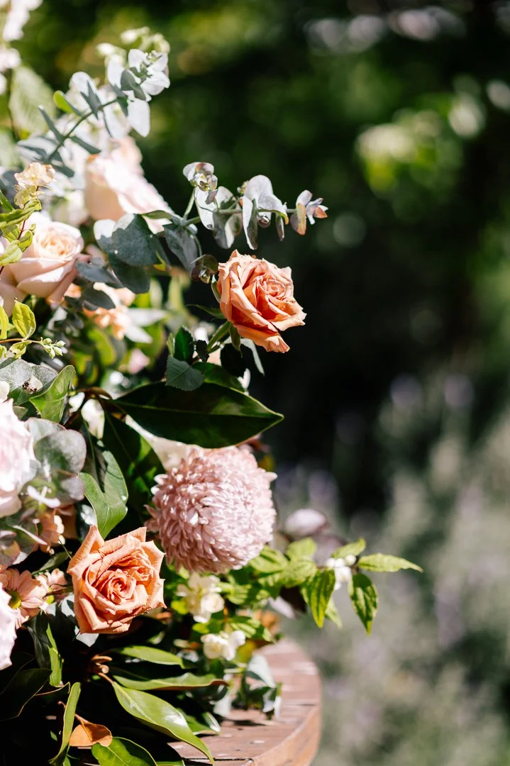 A floral arrangement with peach roses, pink pom-pom flowers, white small blooms, and green eucalyptus leaves on a wooden surface, outdoors with blurred green foliage in the background.