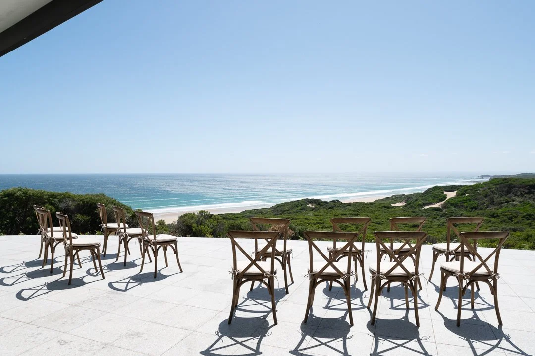 Outdoor patio with eleven chairs overlooking a beach and ocean with greenery and dunes.