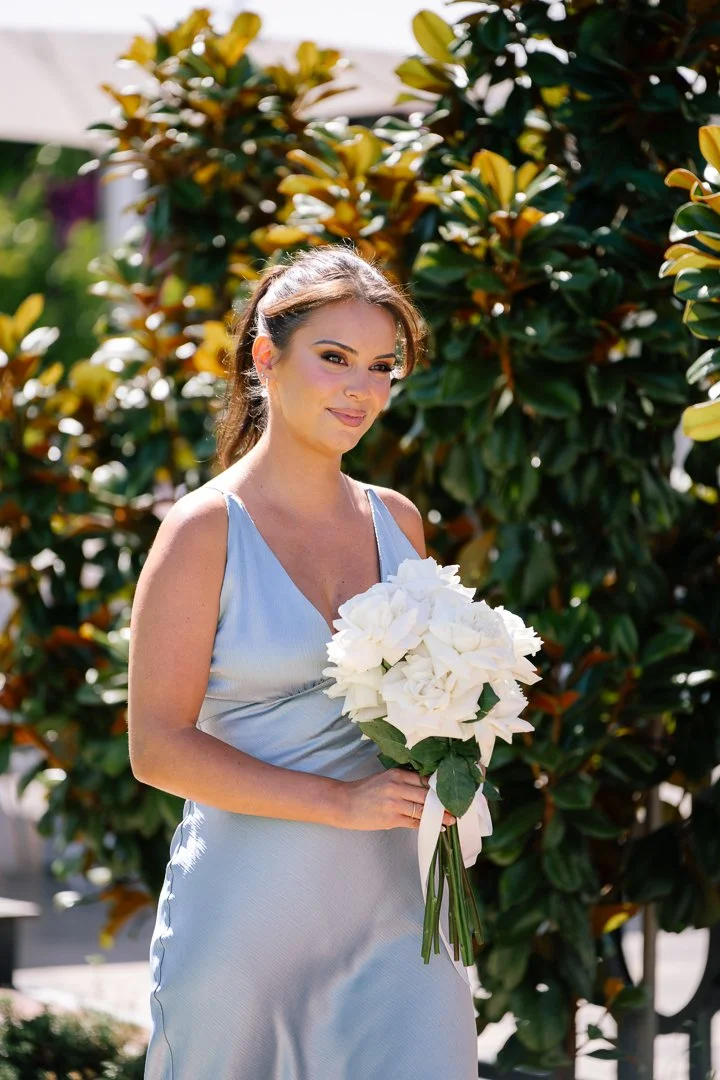 A young woman holding a white bouquet of flowers outdoors, standing in front of leafy greenery, dressed in a light blue satin dress, with sunlight on her face.