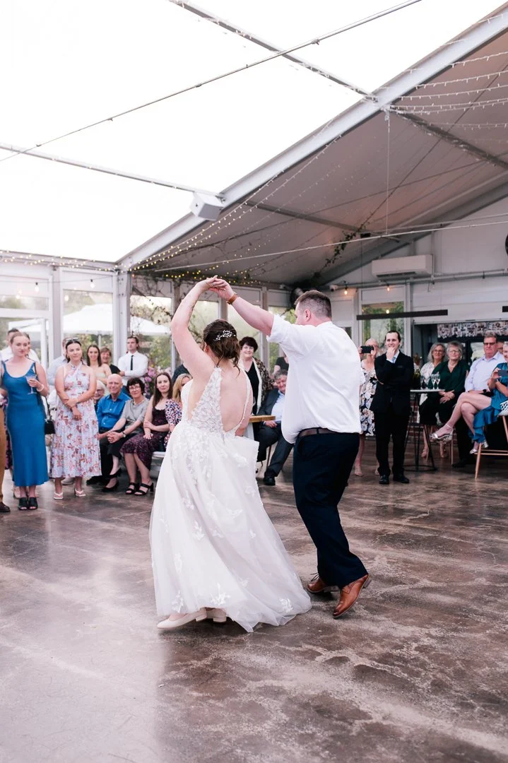 A bride and groom are dancing during their wedding reception with guests watching in a decorated outdoor pavilion.