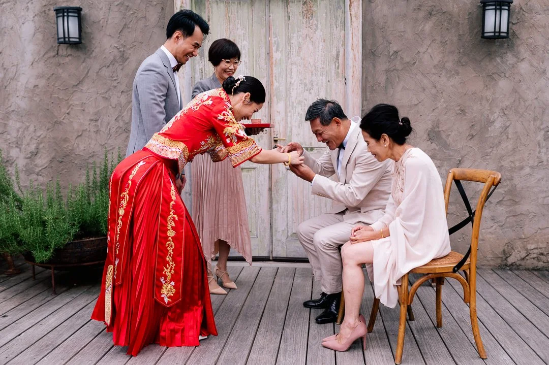 An Asian woman in a red traditional dress presents something to an Asian man in a light-colored suit, who is sitting and smiling. Two women and one man, all dressed formally, stand nearby, watching and smiling. The scene takes place outdoors on a woo