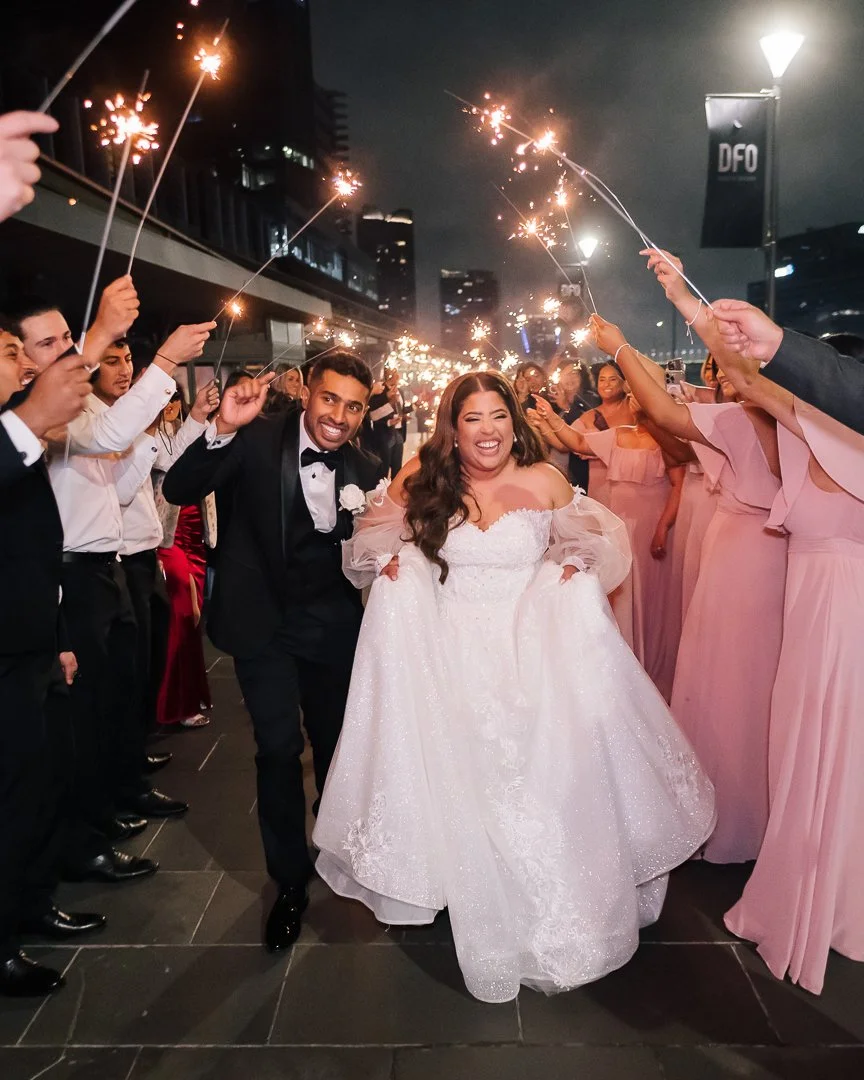 Bride and groom celebrating with sparklers surrounded by wedding guests at night in an urban area.