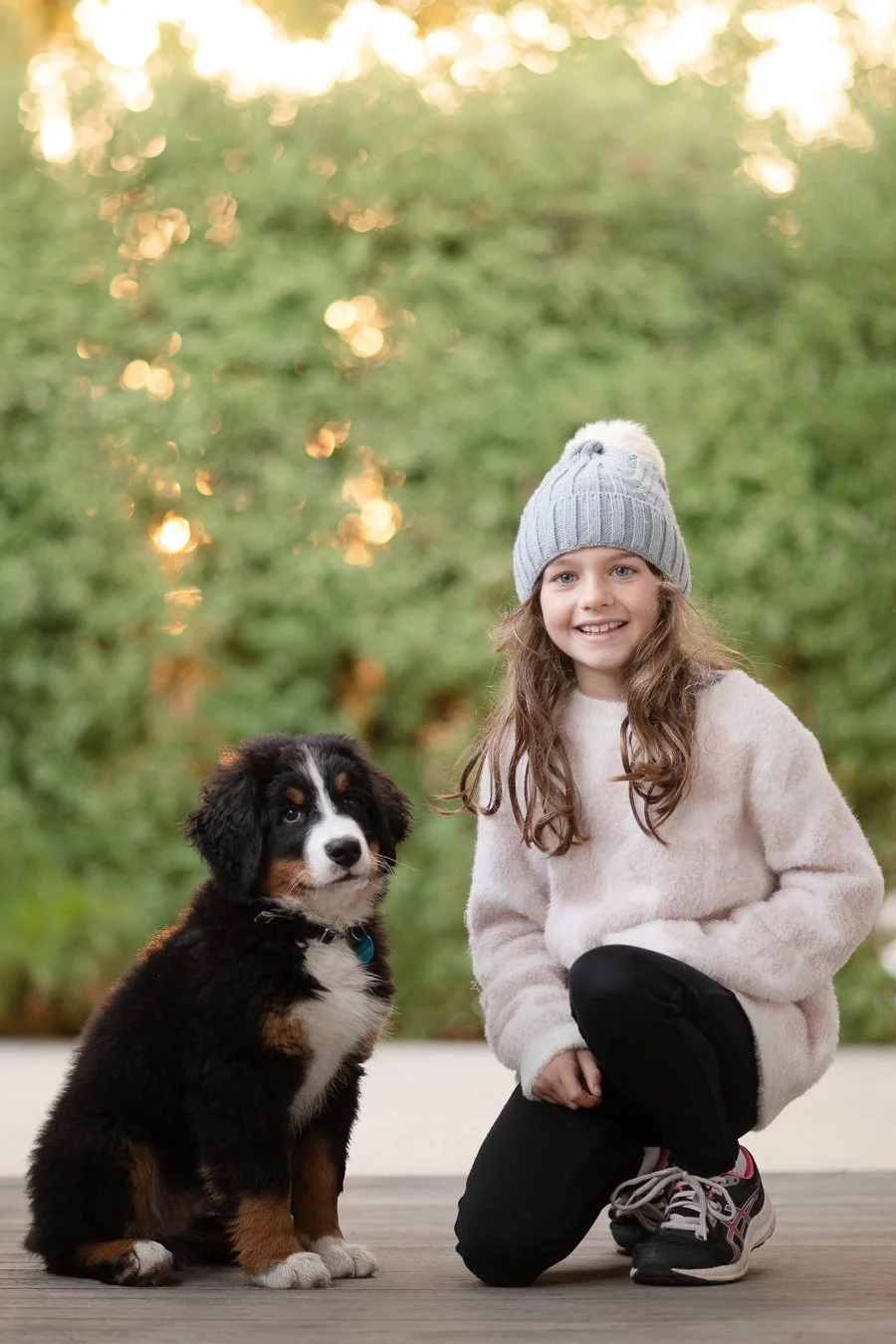 A young girl with long brown hair wearing a gray knit hat, beige sweater, black pants, and sneakers kneeling next to a black, white, and brown puppy in an outdoor setting with blurred green trees in the background.