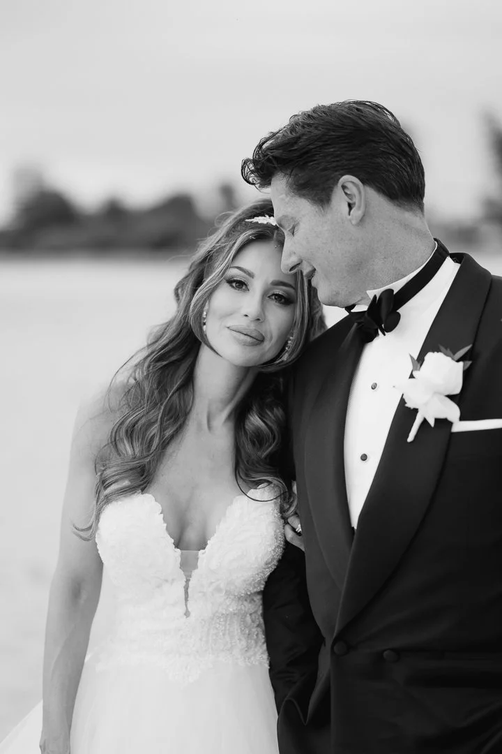 A black and white photo of a bride and groom, with the bride leaning her head on the groom's shoulder, smiling softly. The bride is wearing a strapless wedding dress, and the groom is in a tuxedo with a bow tie and a white boutonniere. The background