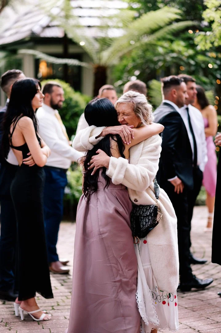 Two women hugging at an outdoor gathering, with people in formal attire in the background.