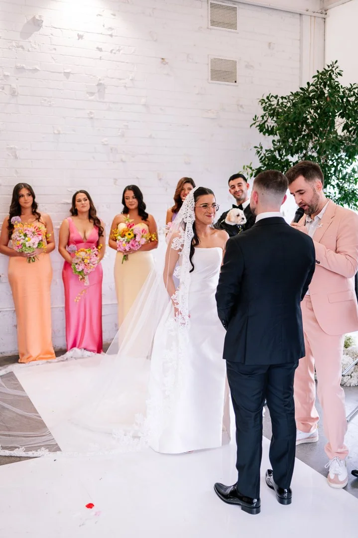 A wedding ceremony with a bride and groom standing in front of officiant, with bridesmaids holding bouquets behind them, against a white brick wall and a large green plant.