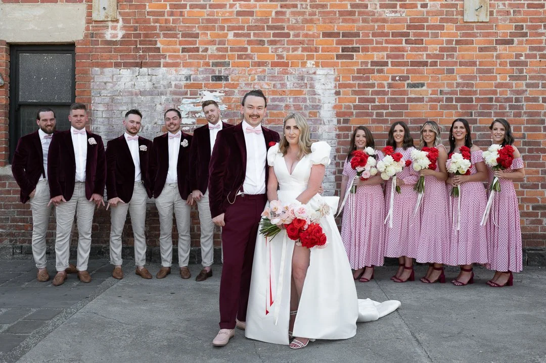 A bride and groom standing with their wedding party against a brick wall, with the bride holding a bouquet of red and white flowers and bridesmaids holding matching bouquets, all dressed in vintage-style attire.