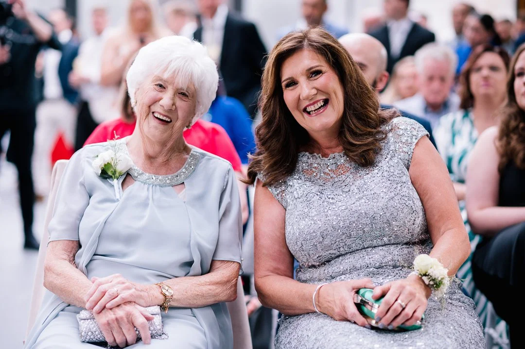 Two women sitting and smiling at a formal event, with other people seated in the background.