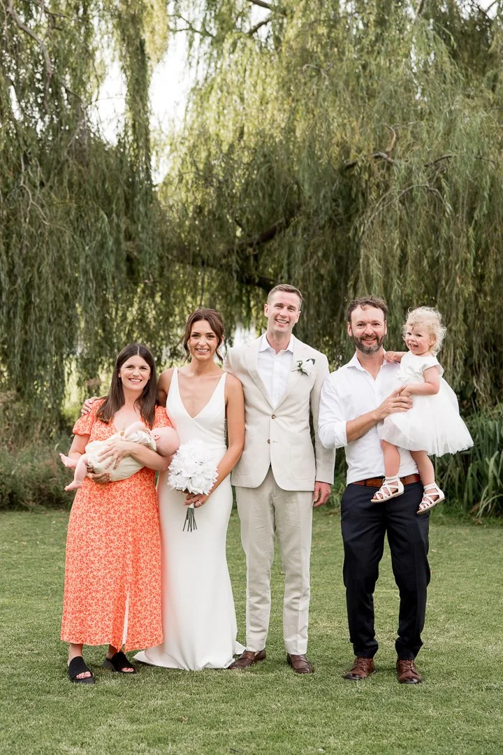 Group of five people standing outdoors on grass, with trees and greenery in the background, celebrating at a wedding or family gathering.