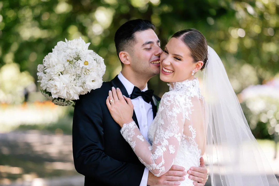 A newlywed couple in wedding attire embracing outdoors, with the bride holding a bouquet of white flowers, both smiling.