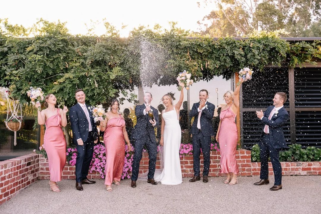 Wedding celebration with the bride and wedding party celebrating outdoors, with champagne pouring, flowers, and smiling. The bride is in the center holding a bouquet, and the wedding party is dressed formally with women in pink dresses and men in sui