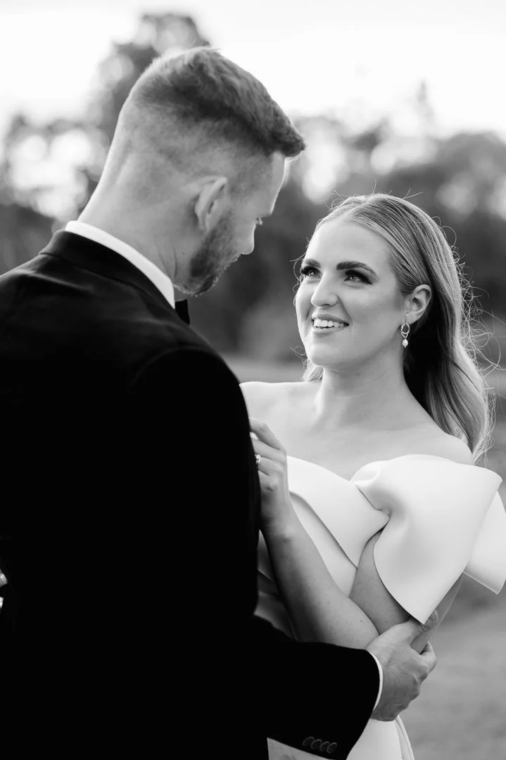 A black-and-white photograph of a couple on their wedding day. The bride is smiling and looking into the groom's eyes, while the groom faces her, holding her gently. The bride is wearing earrings and a dress with a large bow on the shoulder.