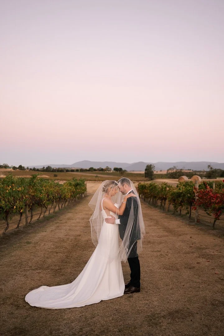 Wedding couple in wedding attire, standing close together in a vineyard at sunset.