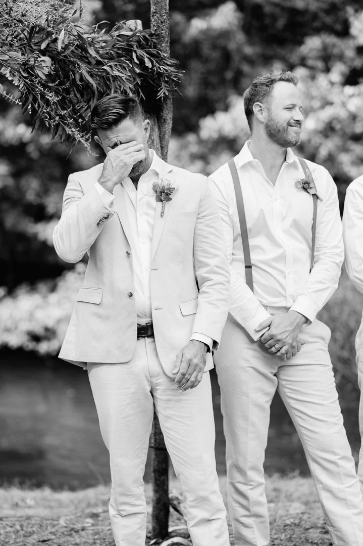 Two men dressed in light-colored suits at an outdoor wedding, one is wiping his face while the other smiles, standing in front of a tree with blurred foliage background.