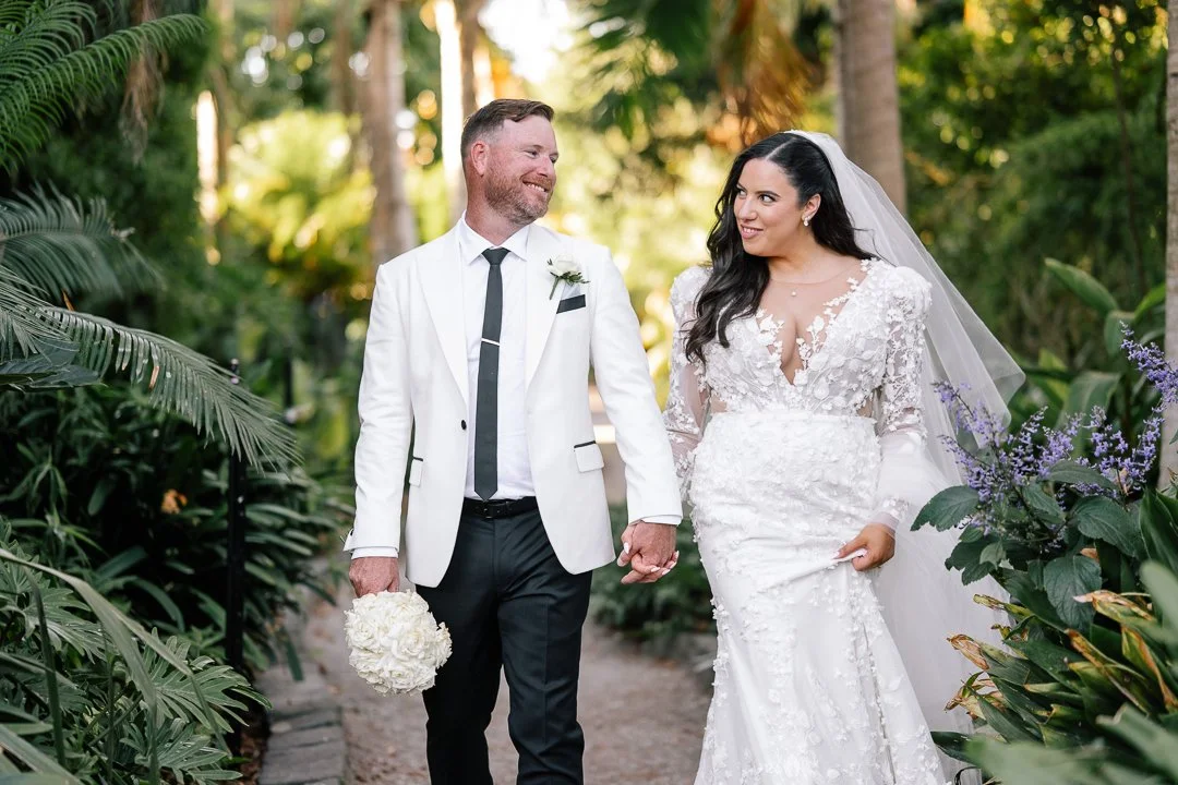 Bride and groom walking hand in hand in a lush garden, smiling at each other, with the bride wearing a white wedding gown and veil, and the groom in a white tuxedo jacket and black pants.