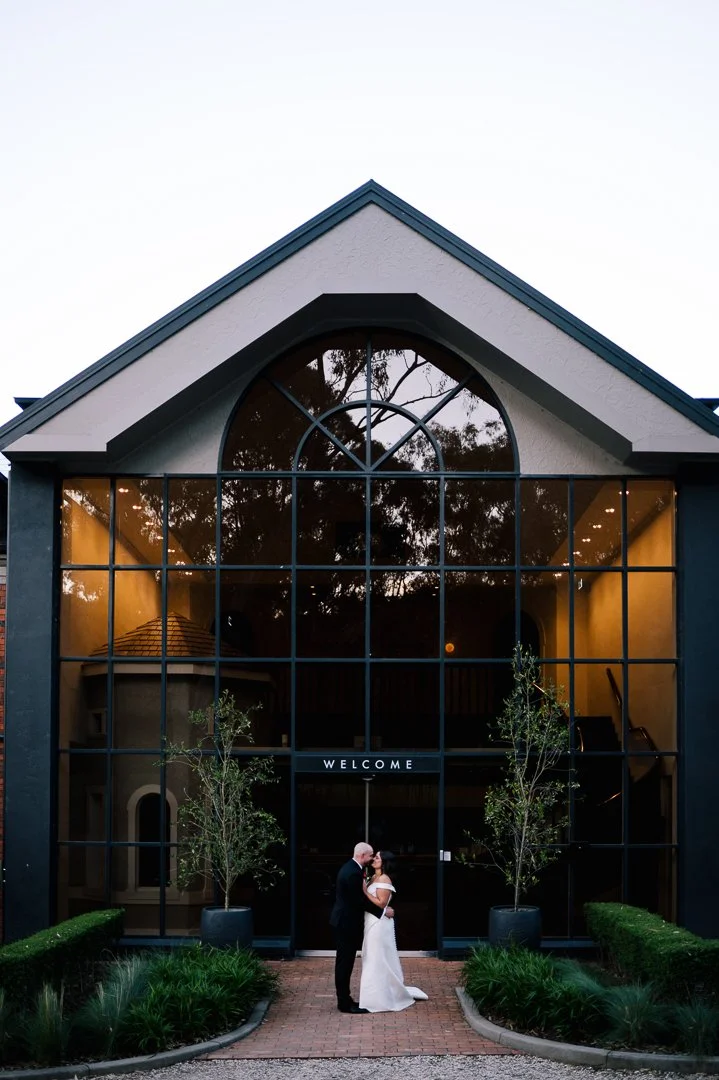 A bride and groom standing in front of a large glass building, holding hands and kissing. The building has a welcoming sign, with trees and landscaped bushes on either side.