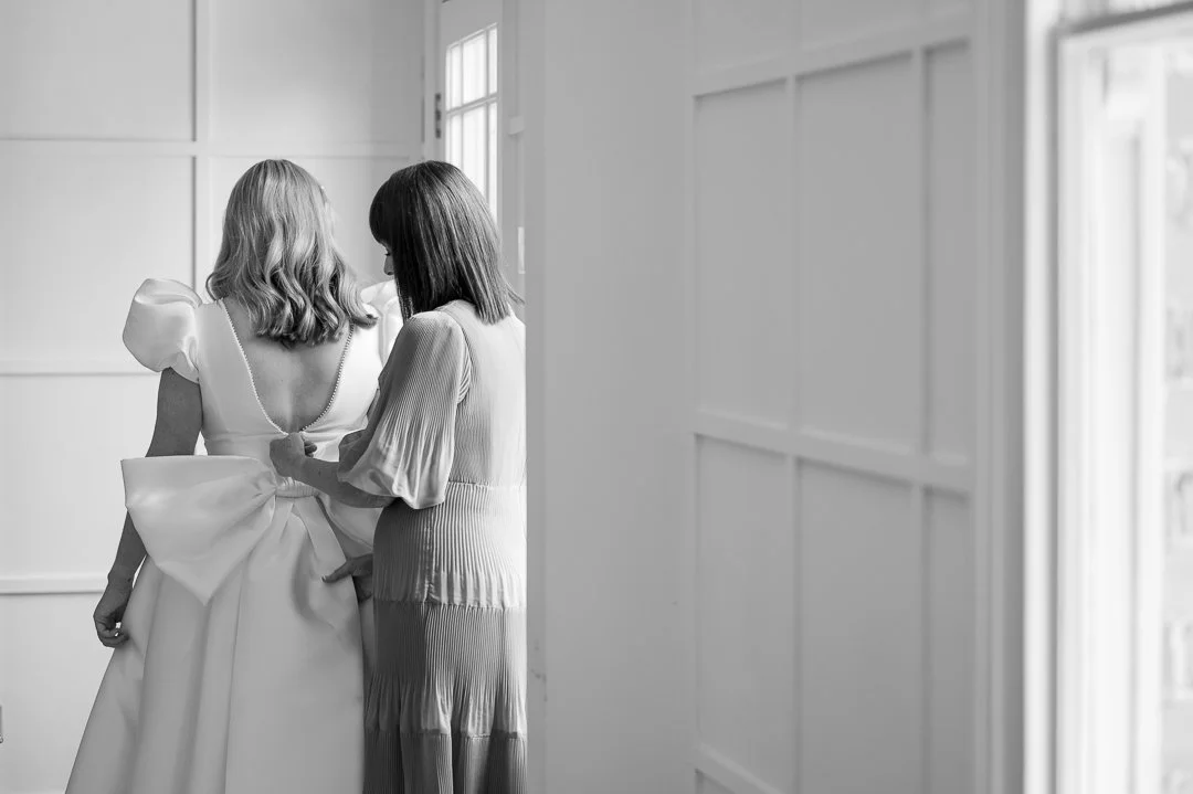 Two women helping a bride with her wedding dress in a bright room.