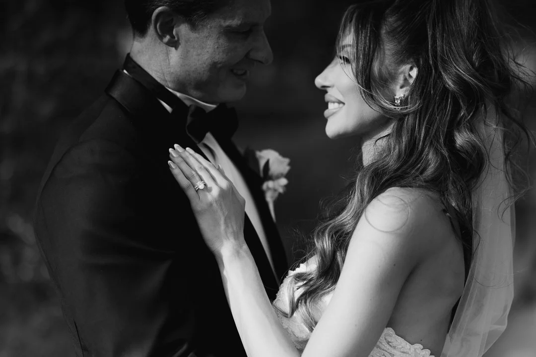 Black and white photo of a bride and groom on their wedding day, smiling and looking into each other's eyes, with the bride touching the groom's suit.