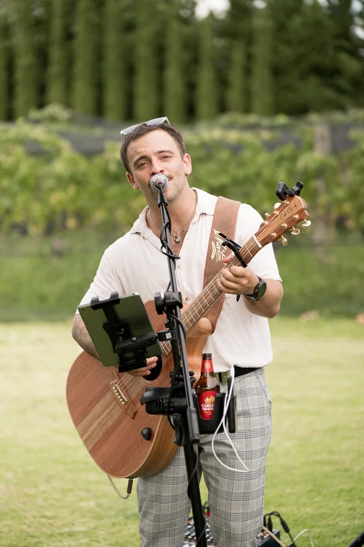 A man performing with a guitar and microphone outdoors in a grassy area with trees in the background. He has sunglasses on his head, is wearing a white shirt and plaid pants, and has a beer bottle in a holder on his waist.