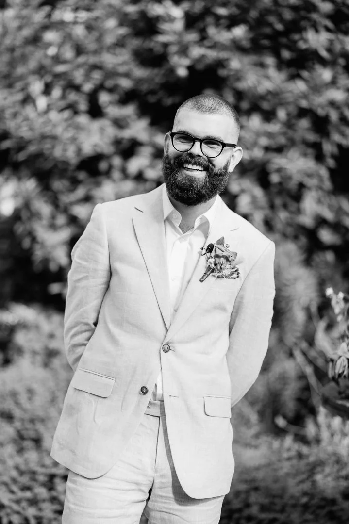 A man with a beard and glasses wearing a light-colored suit and a boutonniere, standing outdoors smiling