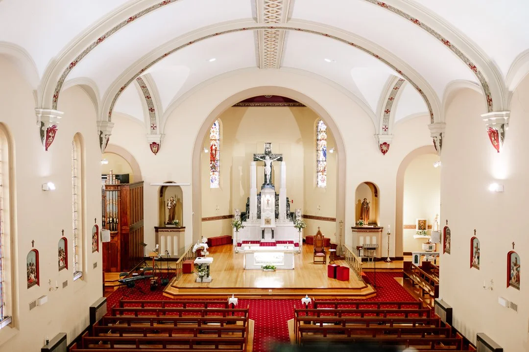 The interior of a church with an altar, crucifix, stained glass windows, wooden pews, and religious statues.