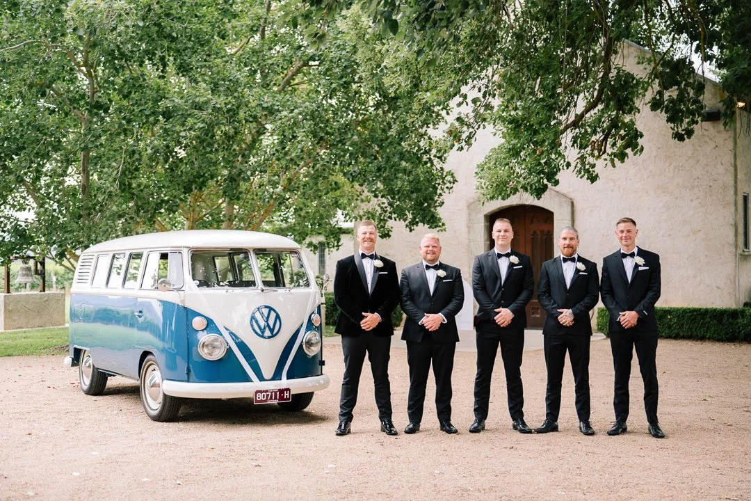 Five men in tuxedos standing outdoors in front of a vintage Volkswagen bus, with a large tree overhead and a building in the background.