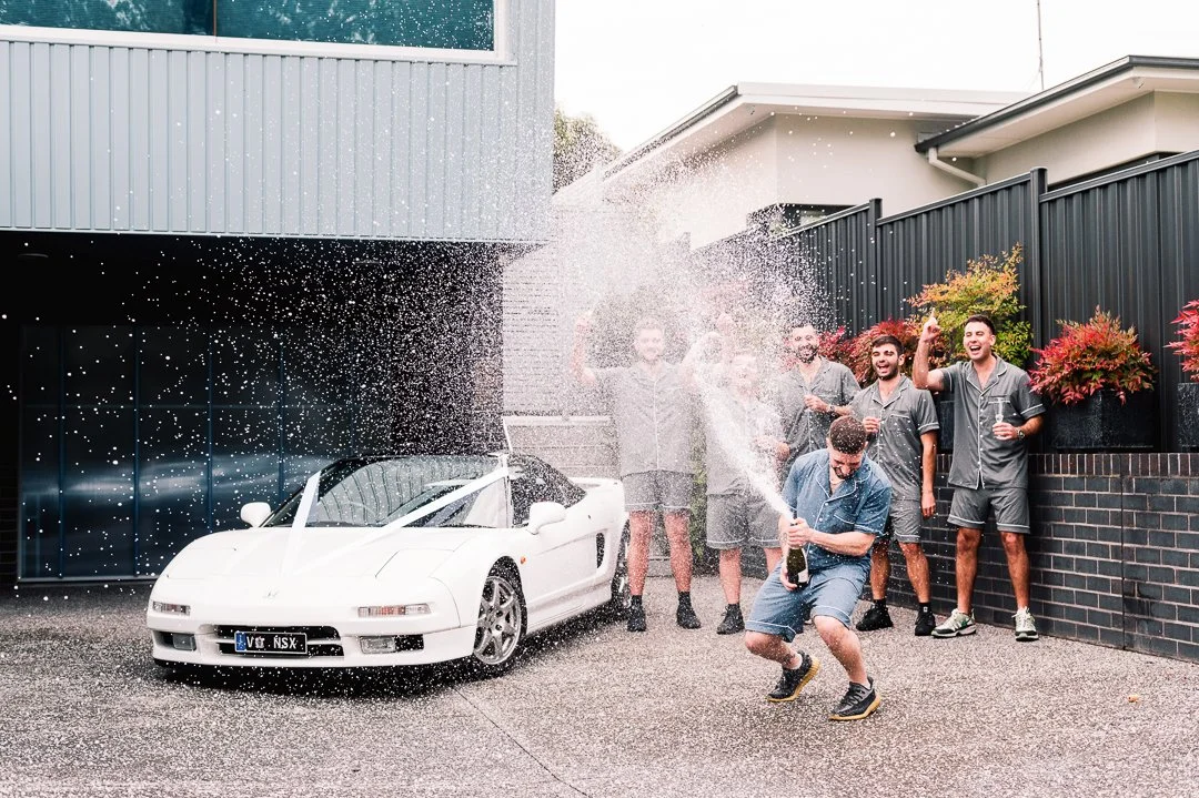 Group of six young men wearing matching pajamas celebrating in front of a white sports car with champagne, some spraying champagne and others holding glasses, outside a modern house with a black fence and colorful plants.