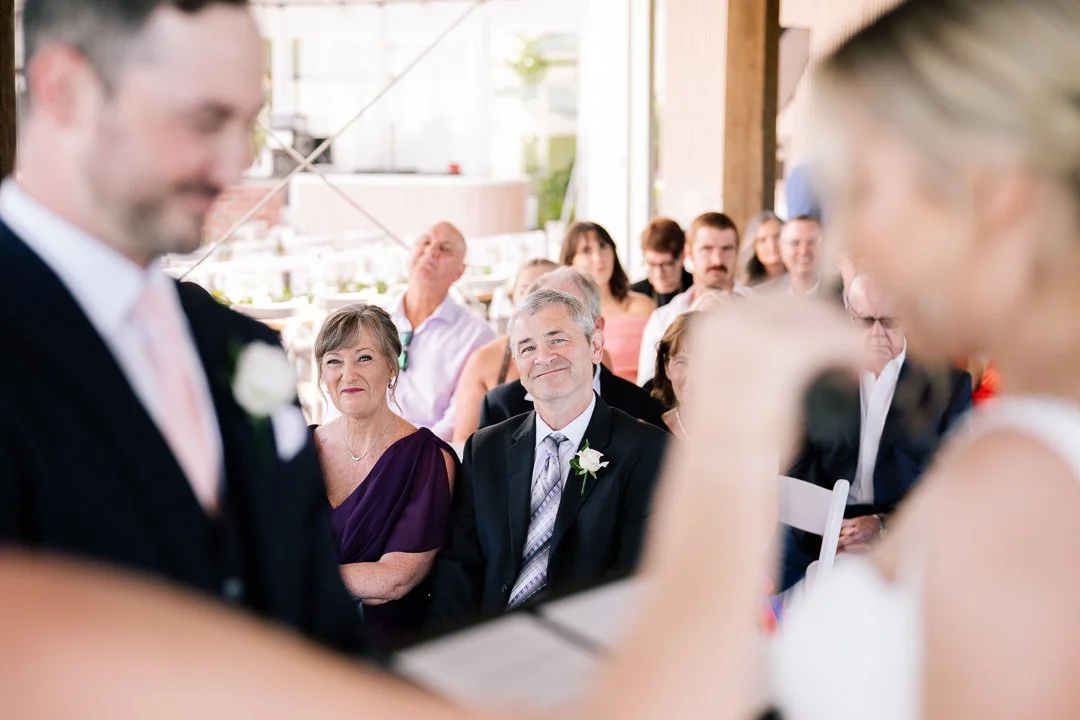 Guests attending a wedding ceremony, with a woman smiling and a man in a suit with a white flower on his lapel looking towards the bride, who is partially visible, in a bright, open indoor setting.