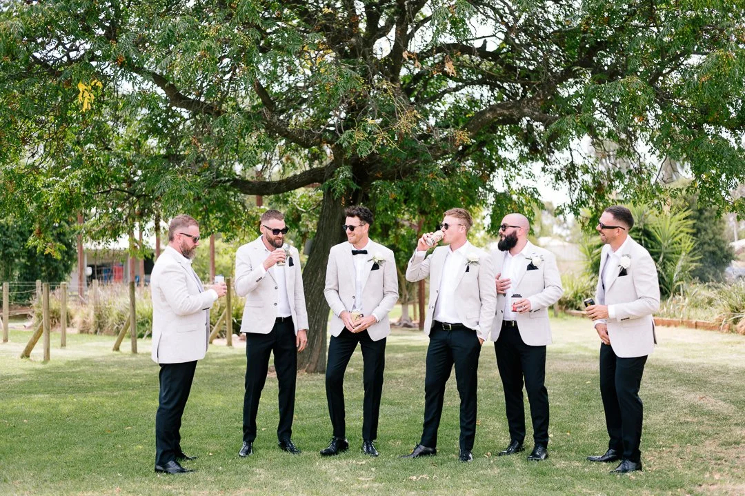 Group of men dressed in tuxedos with bow ties and sunglasses standing on grass under a large tree, drinking and chatting outdoors.
