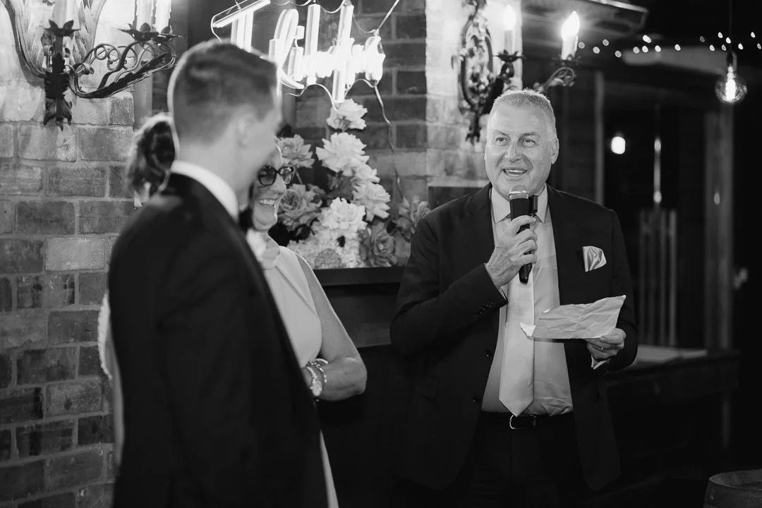 A man giving a speech at a formal event with a microphone and paper, surrounded by others in formal attire, in a decorated indoor setting with flowers and vintage lighting.